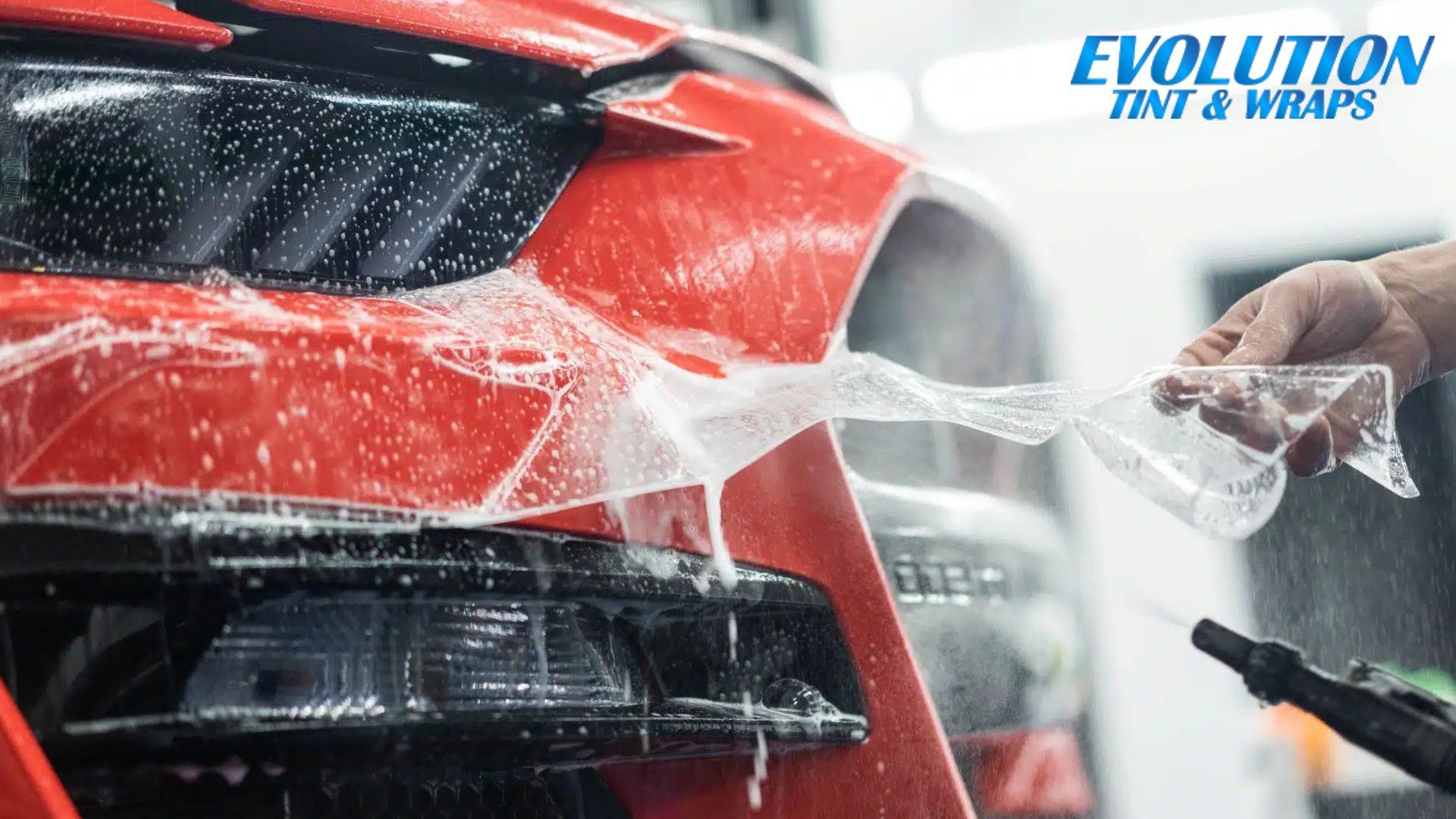 A close-up of a red car's front bumper during a paint protection film application. The film is being stretched and applied while water sprays to ensure proper adhesion