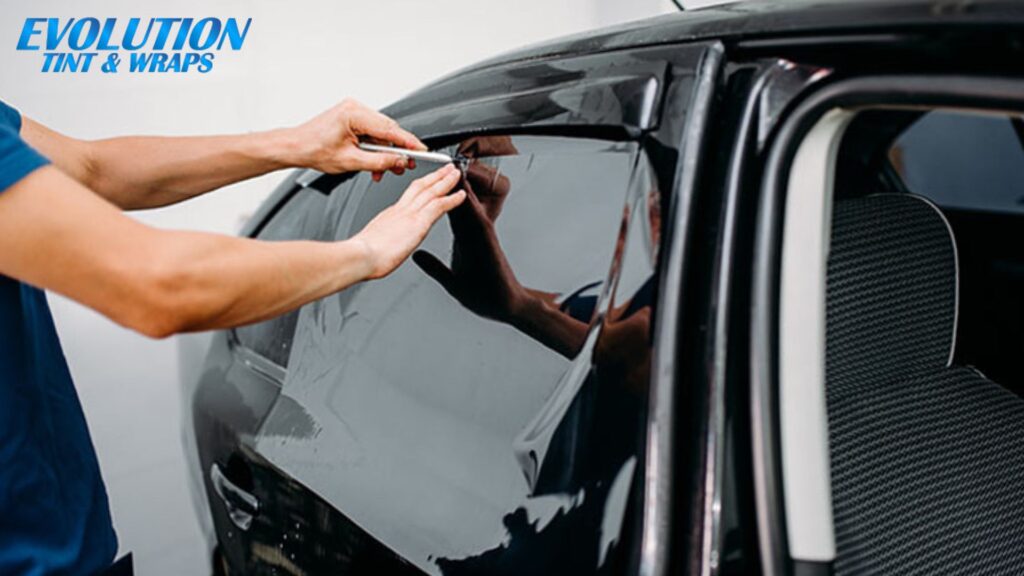 A person trimming excess window tint film on a car's side window, with "Evolution Tint & Wraps" branding visible