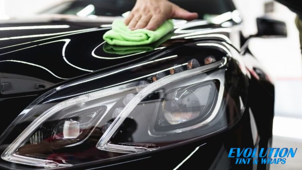 A person wipes the glossy black hood of a car with a bright green microfiber cloth. The car's reflective surface gleams under bright lights