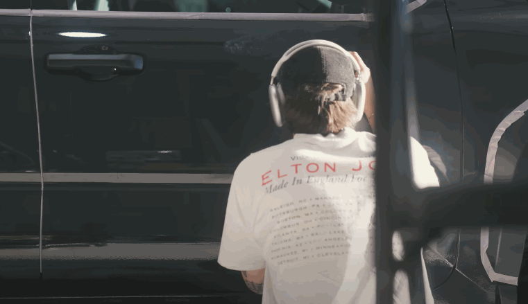 A person wearing headphones and a white shirt featuring Elton John's tour dates works on the side of a black vehicle, polishing the surface with precision