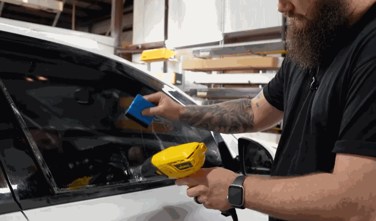 A technician applying window tint to a car window using a heat gun and a smoothing tool for a precise application