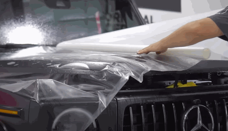 A professional laying a sheet of clear protective film across the hood of a black Mercedes vehicle, preparing it for application as part of a paint protection process