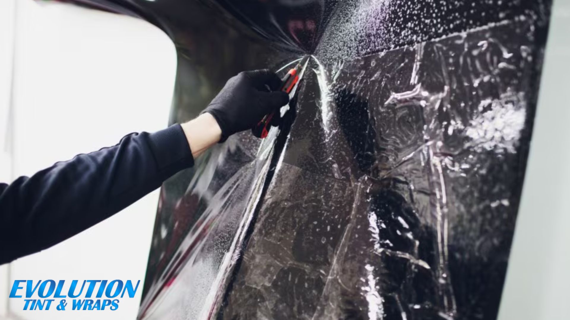 A professional installer carefully trims a protective film on a car’s surface using a precision razor blade, with the "Evolution Tint & Wraps" logo in the corner