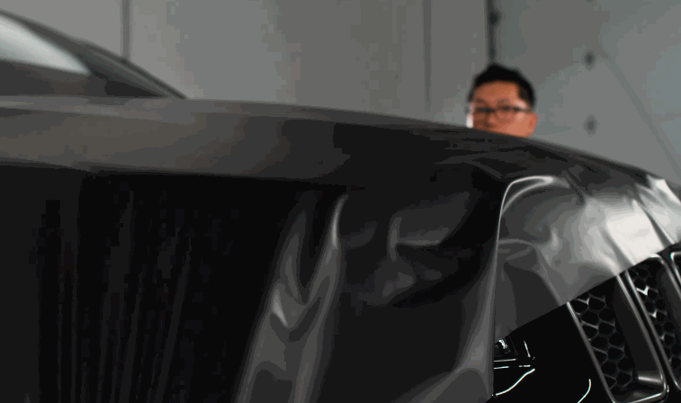 A person carefully aligning a black vinyl wrap on a car hood, ensuring a smooth and professional installation